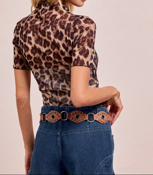 Woman wearing a leopard print top and blue jeans with a belt against a light background