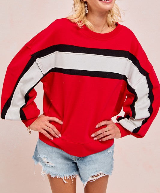 A woman wearing a red oversized sweatshirt with wide black and white stripes across the chest and sleeves, paired with light blue distressed denim shorts, against a solid pale pink background.
