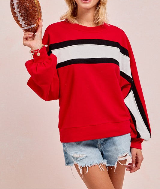 A smiling blonde woman models a red crewneck sweatshirt with bold black and white horizontal stripes across the chest and sleeves. She is holding a glittery brown football prop against a solid light beige background.
