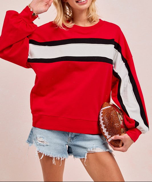 A woman modeling a red crewneck sweatshirt with horizontal black and white stripes across the chest and sleeves, paired with distressed denim shorts and holding a football against a solid light beige background.