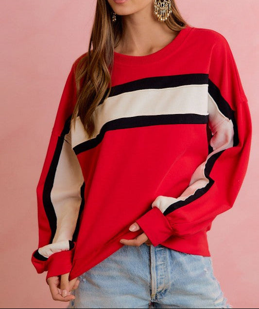 A woman wearing a red long-sleeve sweatshirt with wide black and cream stripes across the chest and sleeves, paired with light blue jeans, set against a solid pink background.