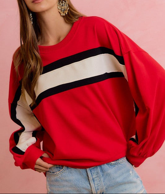 A woman wearing a bright red crewneck sweatshirt with wide black and cream stripes across the chest, paired with light blue jeans, set against a solid pink background.