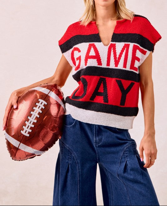 A woman modeling a red, black, and white striped knit sweater with 'GAME DAY' written across the front, paired with wide-leg blue jeans and holding a metallic football-shaped balloon against a light beige background.