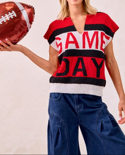 A woman modeling a red, black, and white striped knit sweater with 'GAME DAY' written across the front, paired with wide-leg blue jeans and holding a metallic football-shaped balloon against a light beige background.