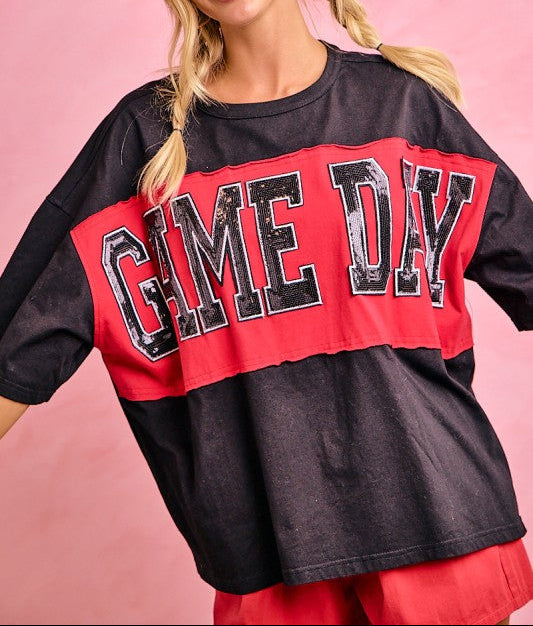 A woman wearing an oversized short-sleeve t-shirt with a color-blocked design in black and red, featuring the text 'GAME DAY' in distressed black letters on the red panel. She is posing against a solid pink background.