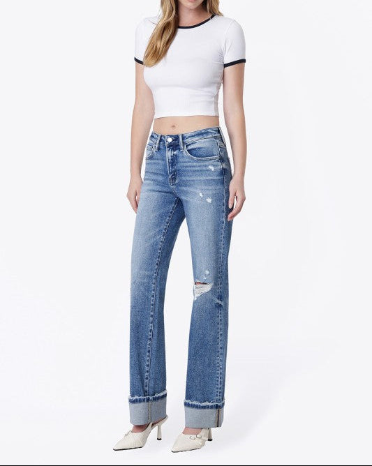 A woman modeling a white short-sleeve crop top with black trim and light-wash blue distressed jeans with cuffed hems, paired with white pointed-toe heels, against a plain white studio background.