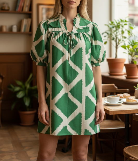 A woman wearing a green and cream geometric patterned mini dress with puff sleeves and a tie-neck detail, standing in a warm, cozy cafe interior.