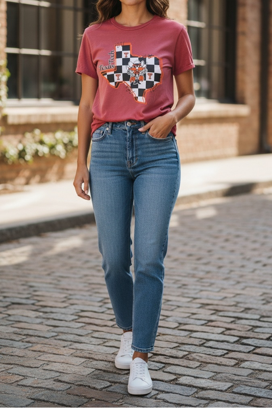 A woman walking on a cobblestone street in front of a brick building, wearing a dusty rose graphic t-shirt with a Texas-shaped patchwork design, blue jeans, and white sneakers.