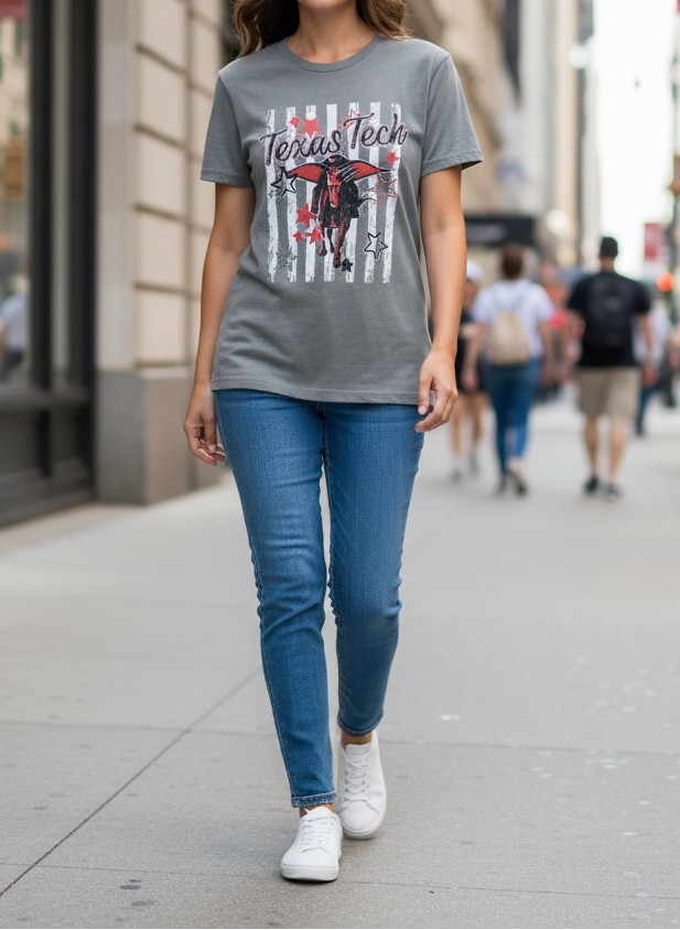 A woman walking on a city sidewalk wearing a heather gray graphic T-shirt with a red, black, and white Chicago Bulls design, blue jeans, and white sneakers.