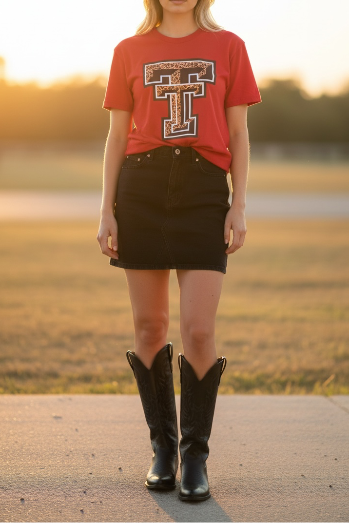 A woman stands outdoors in a field at sunset wearing a red graphic T-shirt with a large collegiate-style letter T, a black mini skirt, and black cowboy boots.